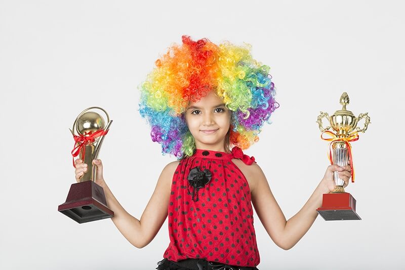 child wearing wig happily posing with trophies
